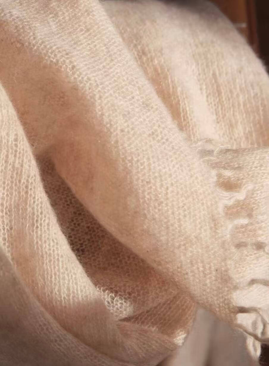 A close-up of a soft, lightweight knit cashmere scarf in a warm, soft pink shade, draped to show its fine texture and fringed edge.