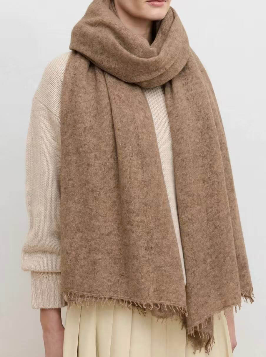 A person wearing a soft, oversized camel-brown cashmere scarf with fringed edges over a cream knit sweater and light beige skirt