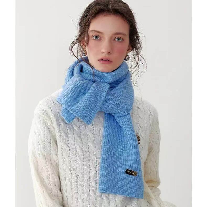 A woman wearing a light blue cashmere ribbed scarf and a white cable knit sweater on a plain background