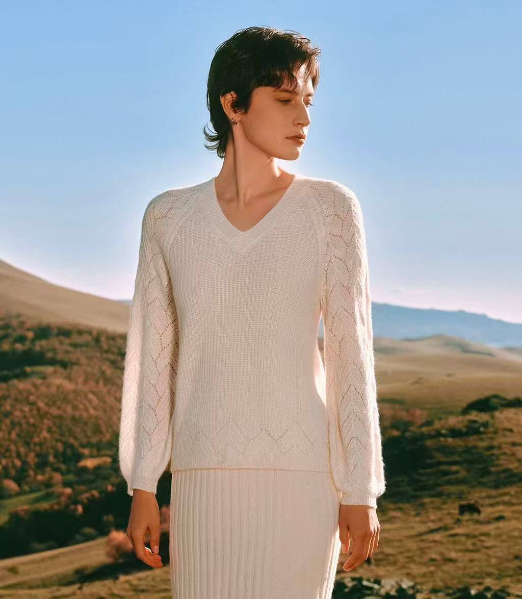 a white cable knit sweater and skirt standing in a scenic outdoor setting with mountains and open fields.
